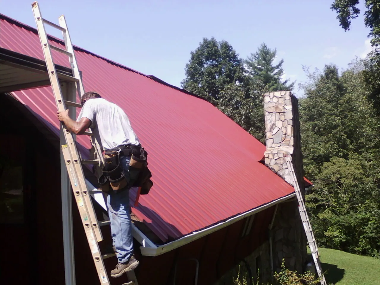 Roofer on a ladder beside a red metal roof and stone chimney — Soffit & Fascia Repair by Roofing Brothers in Showell